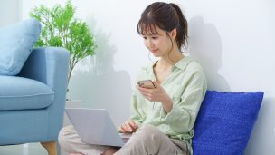 スマホを手にノートパソコンを見る女性